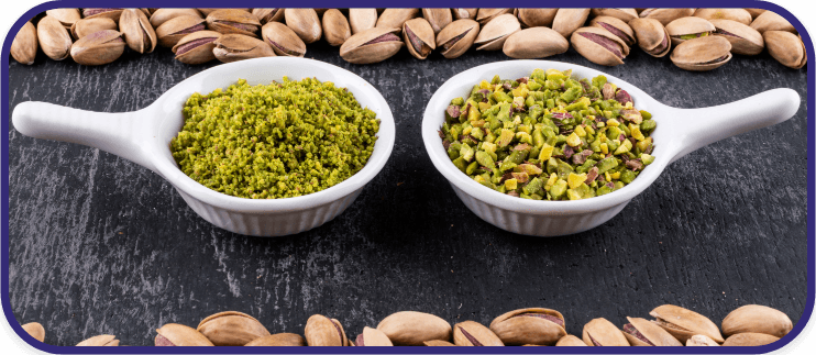 White ceramic bowls filled with ground and chopped pistachios, surrounded by whole pistachio shells on a black stone surface.